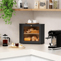 A kitchen setup with a black mini bread box containing pastries, a black espresso machine, a glass French press, a potted plant, and various coffee and tea products stored on a wooden shelf.