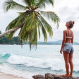 A person stands on a rocky shore, wearing a white bikini top and denim shorts, looking at a leaning palm tree by the ocean. Waves gently roll onto the sandy beach.