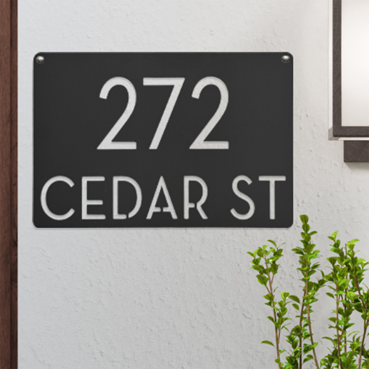 Black and white address plaque displaying \“272 Cedar St\“ with a modern design, attached to a light-colored wall, adorned by greenery.