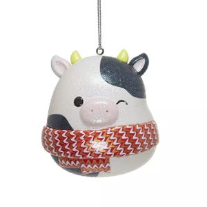 A cute cow-shaped ornament with a winking face, wearing a red and white zigzag-patterned scarf. It features black spots and small horns, ideal for festive decorations.