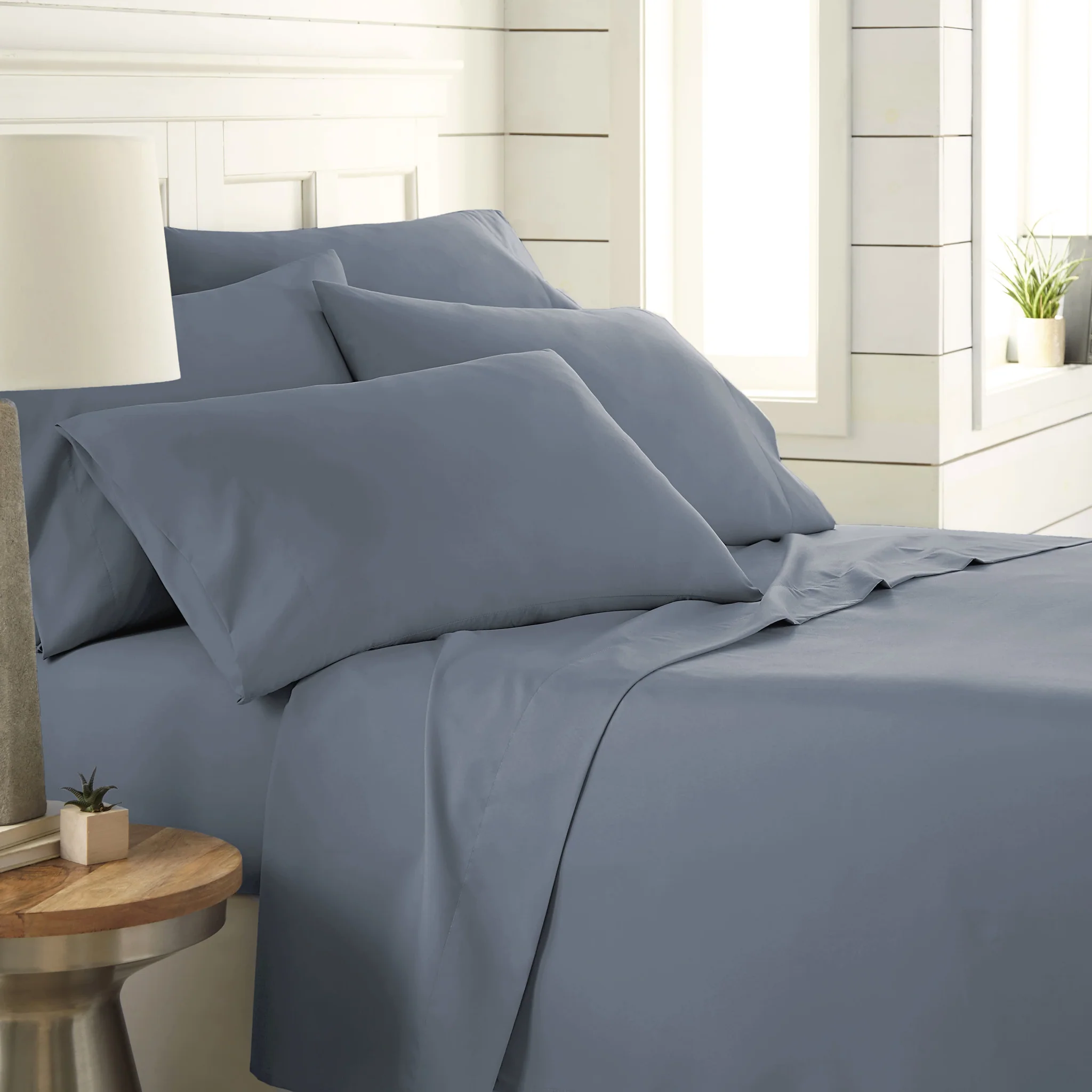 A neatly arranged bed features light blue sheets and pillowcases, accompanied by a small plant on a bedside table in a bright, modern bedroom setting.