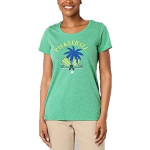 A woman wears a green T-shirt with a \“Pickleball\“ graphic featuring palm trees and paddles, paired with beige pants.