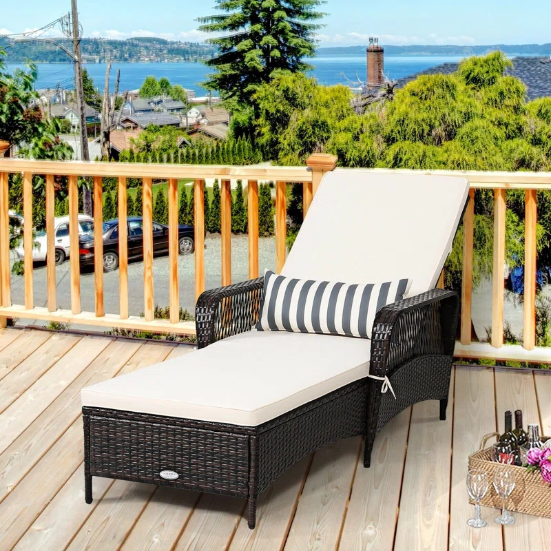Outdoor steel chaise lounge with dark wicker frame, beige cushion, and a black-and-white striped pillow, set on a wooden deck overlooking a scenic view.