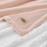 A soft pink UGG blanket with a logo patch on a fluffy white surface.