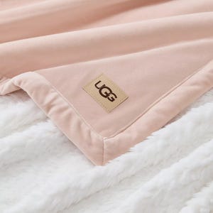A soft pink UGG blanket with a logo patch on a fluffy white surface.