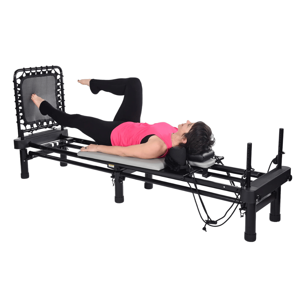 A person is exercising on a Pilates reformer machine, performing a leg exercise while lying on their back, with one foot on a rebounder. The apparatus features adjustable straps and a cushioned platform for support.