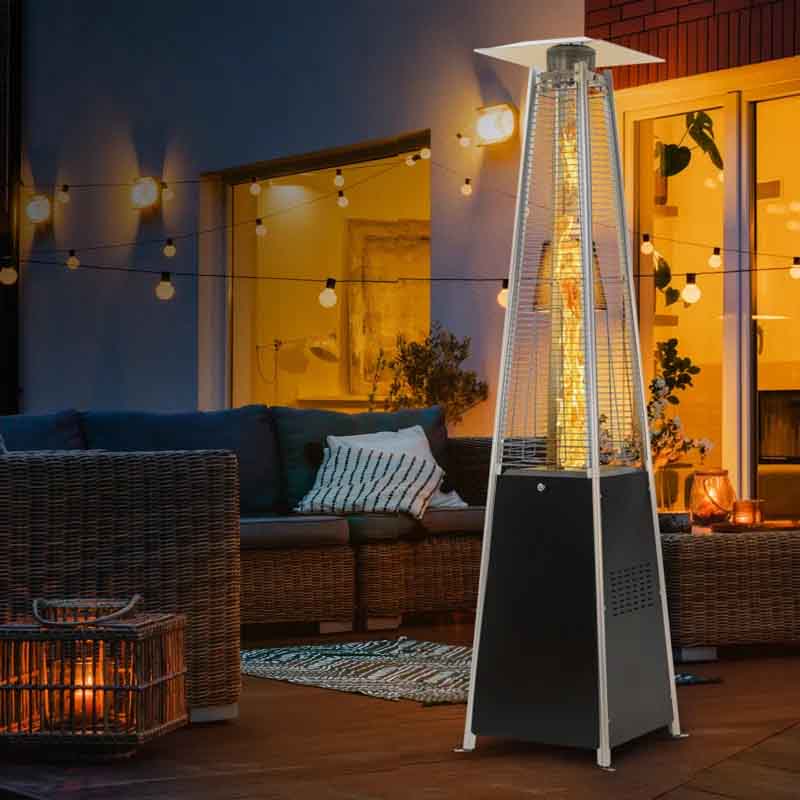 Outdoor patio heater with a modern design, set on a cozy deck with wicker furniture, cushions, string lights, and a warm indoor ambiance visible through glass doors.