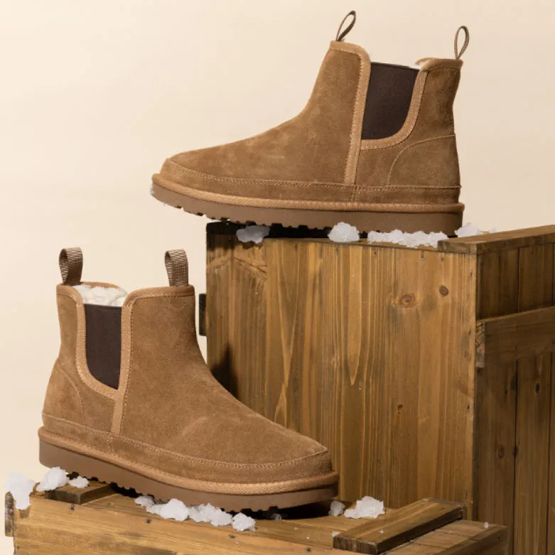 Tan suede ankle boots with pull tabs and dark elastic side panels, resting on wooden crates with scattered faux snow.