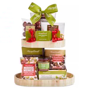 A tiered gift basket features Harry & David items including Moose Munch, Mediterranean Herb Crackers, dipping pretzels, sweet and spicy snack mix, red candy, dark and milk chocolate truffles, sesame sticks, and classic caramel sauce, accented with a green bow.