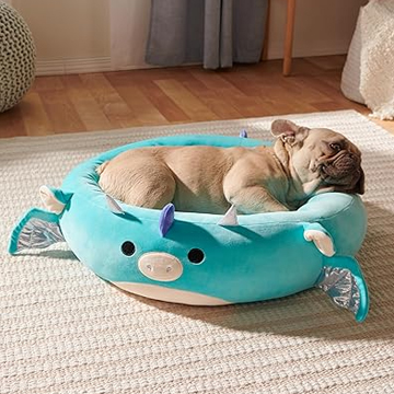 A dog is lying in a plush pet bed designed to resemble a cute, stylized blue dragon with details like little wings and horns.