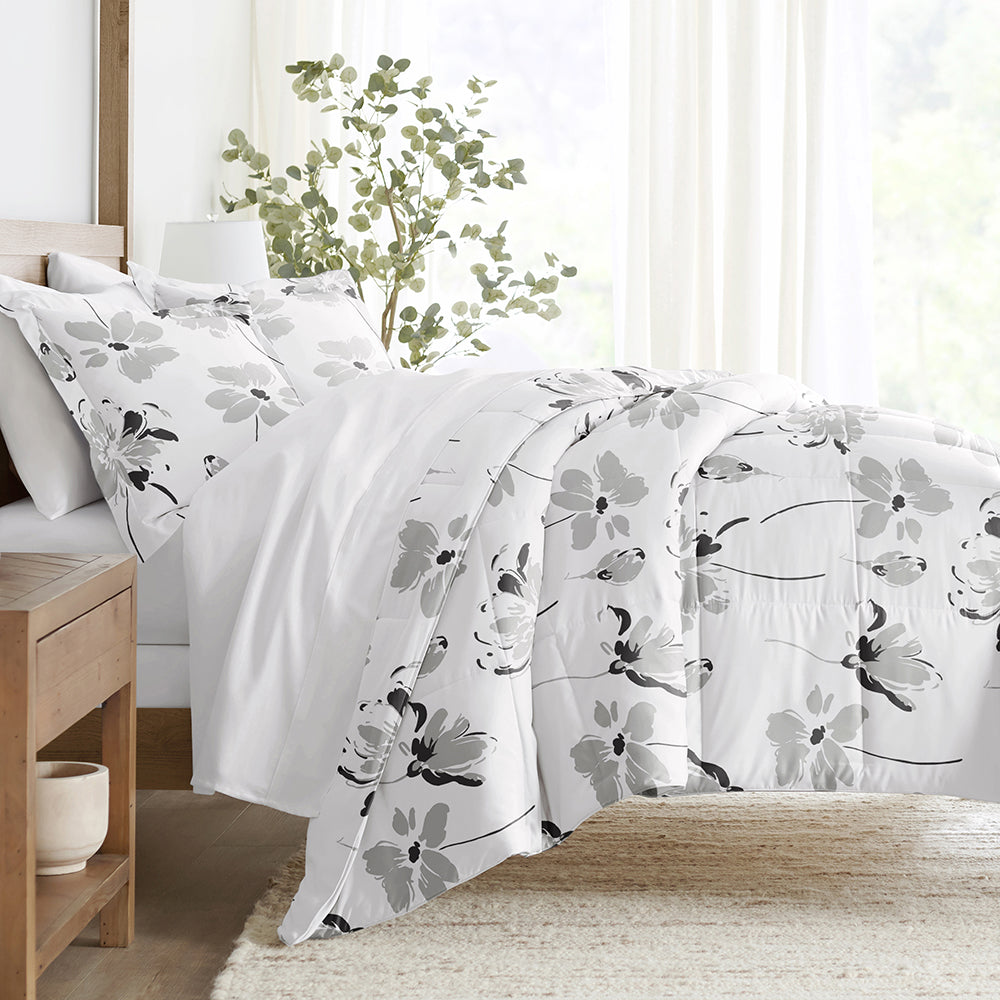A floral-patterned comforter set featuring black, grey, and white flowers on a white background. The bed is complemented with matching shams and a light wooden side table, placed in a bright room with plants.