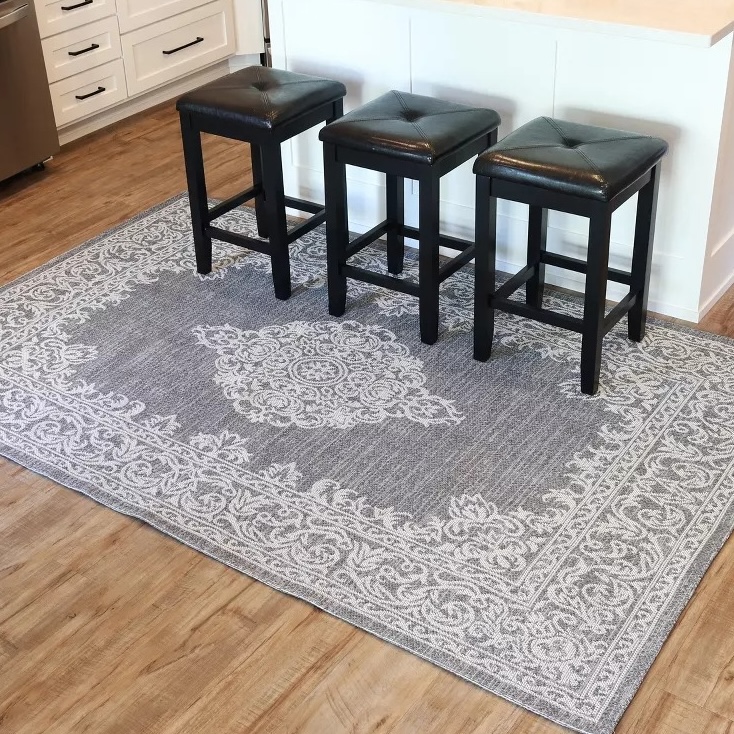 A stylish gray and white decorative rug with a central medallion pattern is placed beneath three black bar stools with cushioned seats in a kitchen with light wood flooring and white cabinetry.