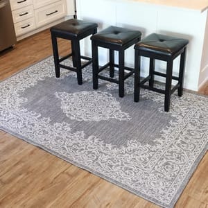 A stylish gray and white decorative rug with a central medallion pattern is placed beneath three black bar stools with cushioned seats in a kitchen with light wood flooring and white cabinetry.
