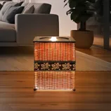A woven rectangular lamp with floral patterns emits a warm glow, placed in a modern living room setting with a sofa, rug, and potted plant.