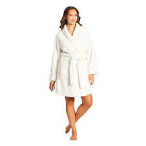 A woman in a white plush robe with a shawl collar, long sleeves, and two front pockets, cinched at the waist with a matching belt.