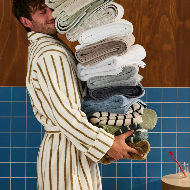 A person in a striped bathrobe holds a large stack of rolled towels of various colors, with a tiled blue wall and a wooden surface in the background.