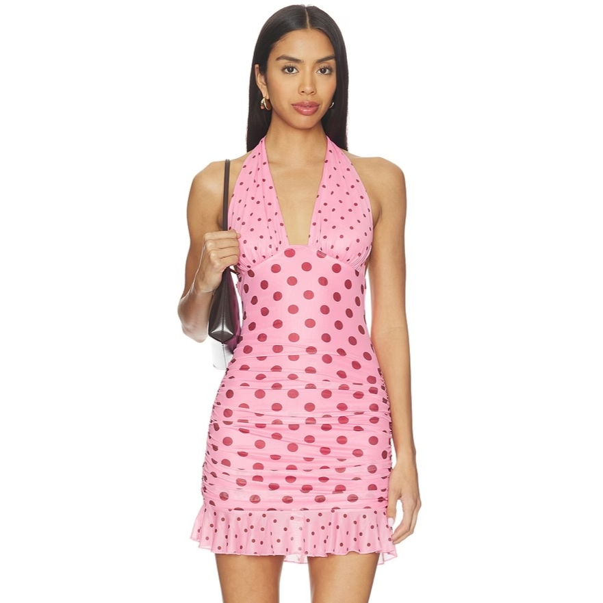 A woman is wearing a pink polka dot halter-neck mini dress with ruffled hem, carrying a black handbag.