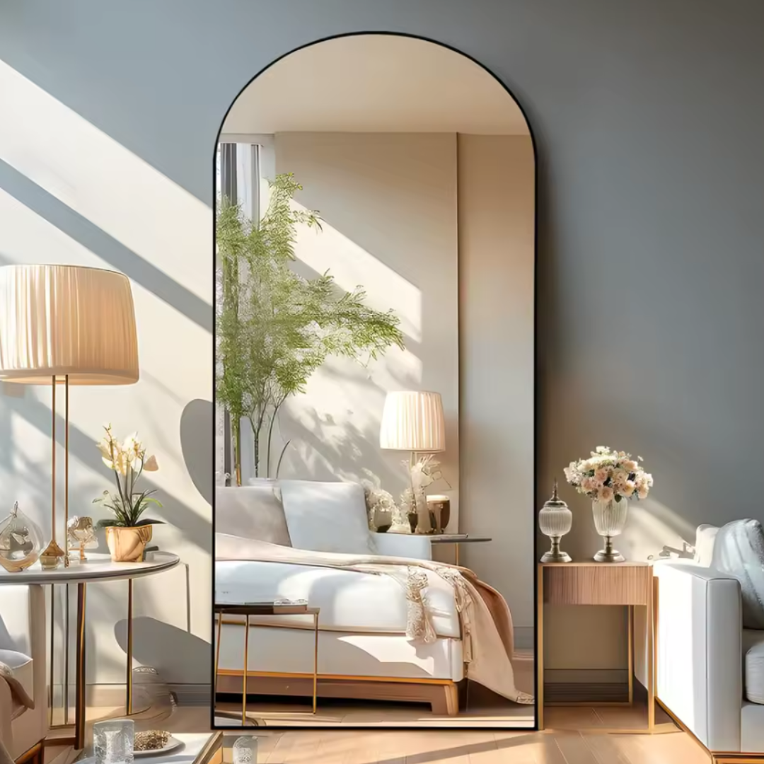 The oversized full-body mirror features an elegant arched design with a thin black frame. It reflects a sophisticated living space with modern decor, including a lamp, side table, and soft furnishings, creating an expansive and stylish aesthetic.