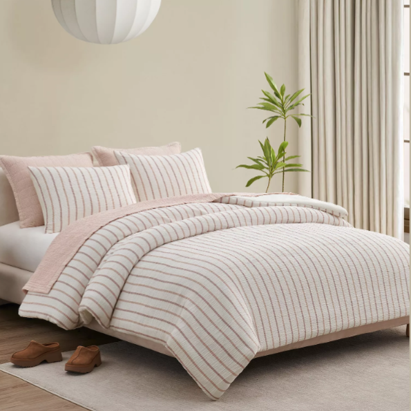 A neatly made bed features striped bedding in soft tones, complemented by matching pillows. The room includes a round pendant light, a potted plant, beige curtains, and brown shoes on a light rug.