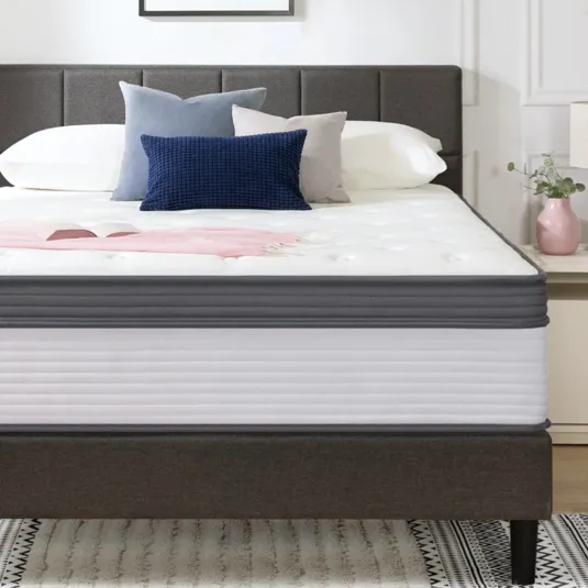 A modern bedroom setup features a bed with a plush mattress, several decorative pillows, and minimalistic bedside tables with lamps. A simple abstract painting adorns the wall above.
