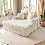 A large, plush, white modular sofa with ribbed texture, accompanied by matching pillows, is situated on a light round rug, surrounded by minimalistic decor and indoor plants by a window.