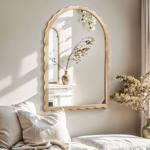 A scalloped-edge mirror hangs on a wall, reflecting a vase with flowers on a mantel. Cream-colored bedding and curtains surround the scene, adding a soft, neutral feel.