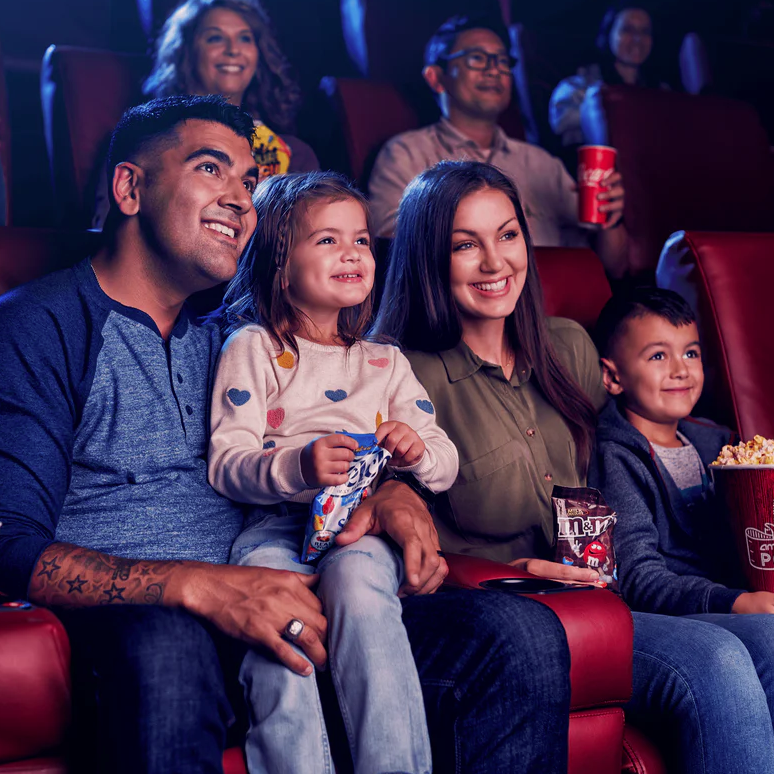 A family enjoys snacks while watching a movie, holding Skittles, M&M's, and a large popcorn in a theater setting.