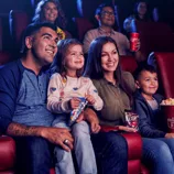 A family enjoys snacks while watching a movie, holding Skittles, M&M's, and a large popcorn in a theater setting.