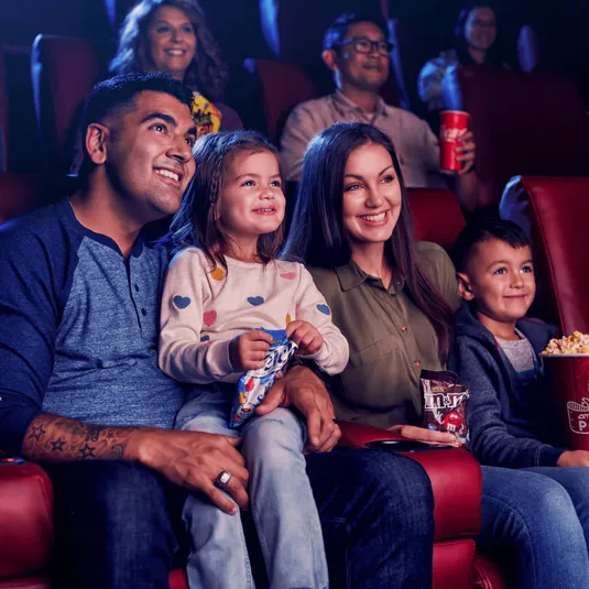 A family enjoys snacks while watching a movie, holding Skittles, M&M's, and a large popcorn in a theater setting.