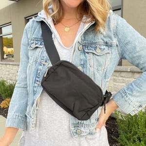 A person is wearing a light blue denim jacket over a gray hoodie and a black crossbody waist pack across the chest. The pack features an adjustable strap and a compact, rectangular shape.