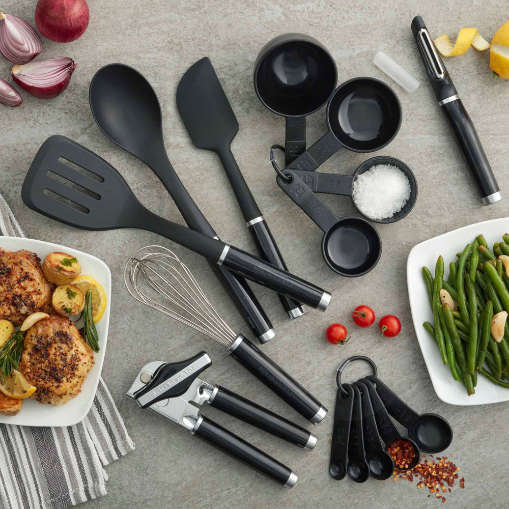 Assorted kitchen utensils including measuring cups, a whisk, spatula, tongs, and a can opener, laid out with prepared food dishes.