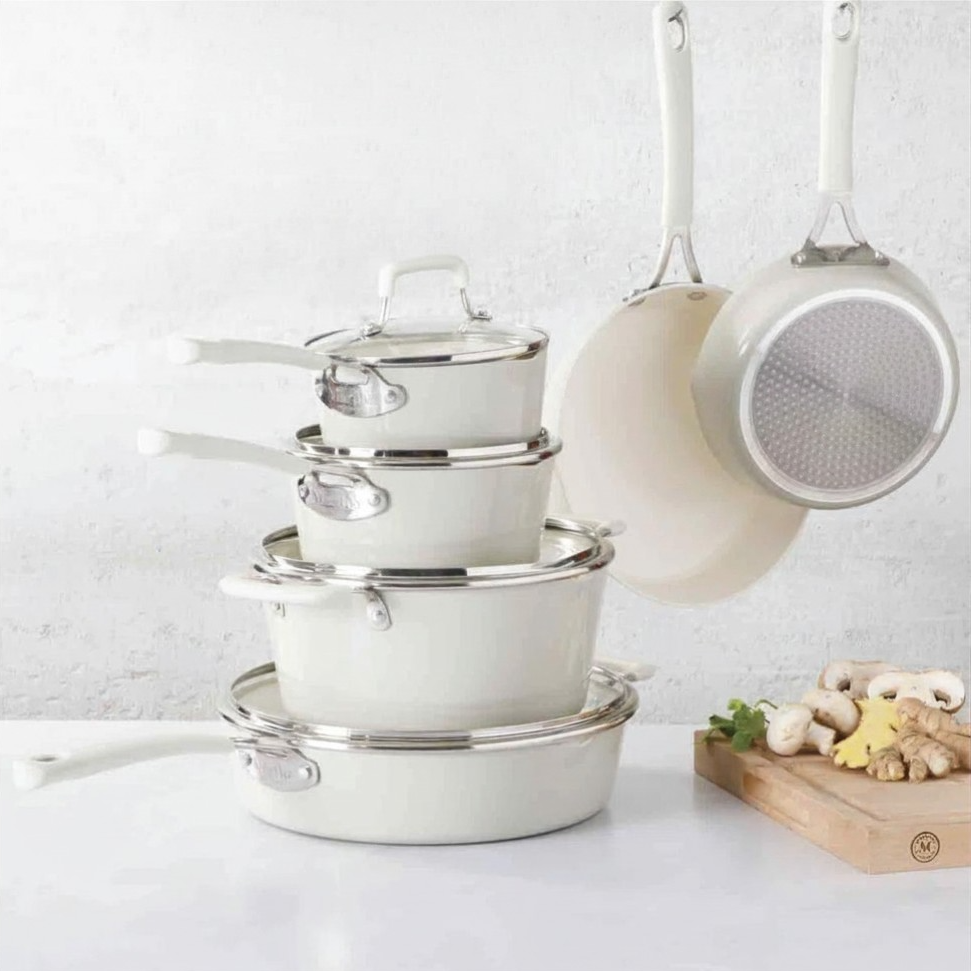 A stack of white saucepans with lids and hanging pots is arranged on a kitchen countertop, accompanied by a cutting board with mushrooms and ginger.