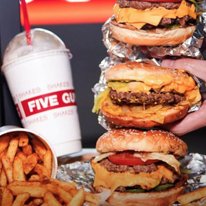 A stack of cheeseburgers and a cup of fries are displayed next to a milkshake with a \“FIVE GUYS\“ logo.