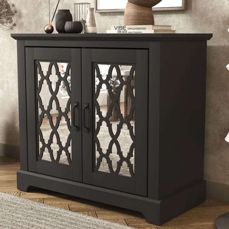 Black accent cabinet with mirrored doors featuring an intricate lattice design, two central handles, and decorative items on top, set on a wooden floor.