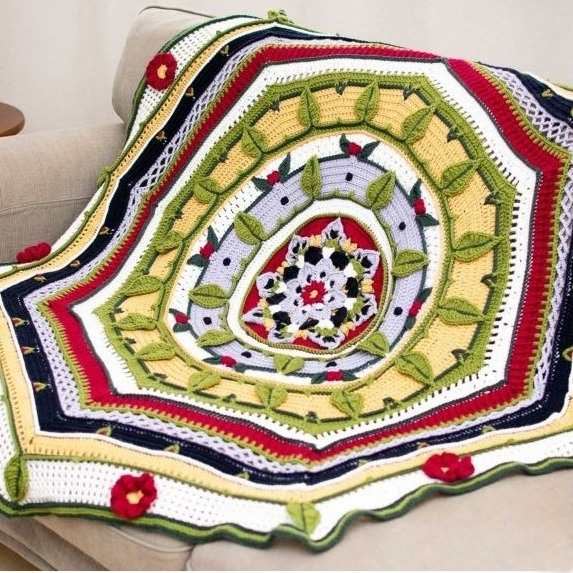 A colorful crocheted blanket with intricate patterns and floral designs is draped over a beige sofa. The blanket features red, green, yellow, and black accents with raised leaf motifs.