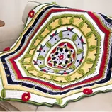 A colorful crocheted blanket with intricate patterns and floral designs is draped over a beige sofa. The blanket features red, green, yellow, and black accents with raised leaf motifs.