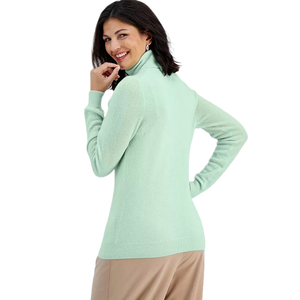 A woman wears a light green cashmere turtleneck sweater from Macy's, paired with beige pants. The sweater has a smooth texture and a fitted silhouette.