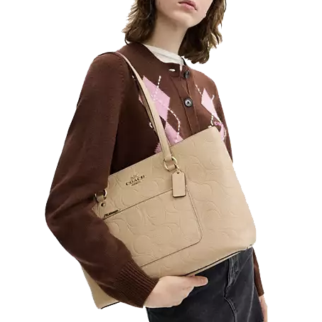 A woman holds a beige Coach shoulder bag with embossed patterns, wearing a brown sweater with pink and white diamond designs.