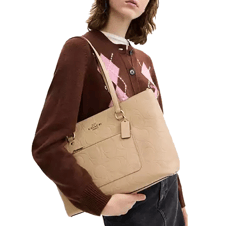 A woman holds a beige Coach shoulder bag with embossed patterns, wearing a brown sweater with pink and white diamond designs.