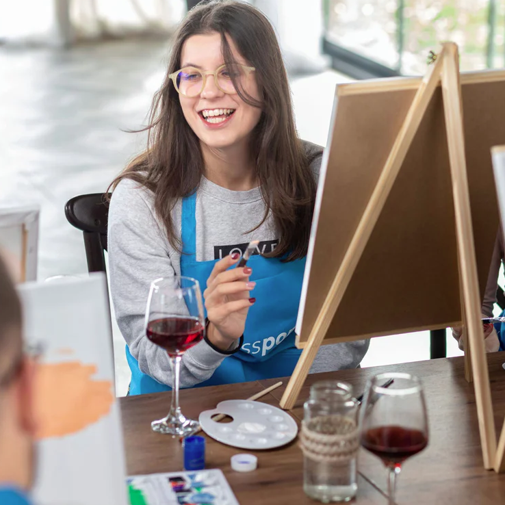 A person is painting on a canvas, wearing a blue apron. There's a glass of red wine, a paint palette, brushes, and art supplies on the table.