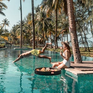 A person dives into a pool while another eats from a floating breakfast tray nearby. Palm trees line the background in a tropical setting.
