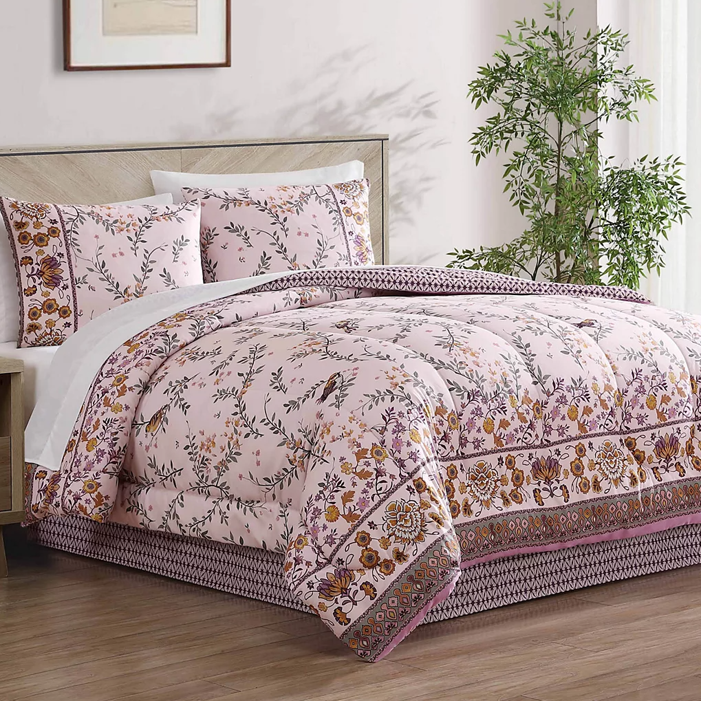 A floral-patterned bedding set including a comforter and pillow shams on a bed, with pink and floral prints. A tall green plant stands to the side in a softly lit room.