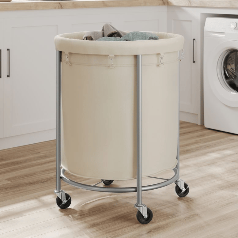 A rolling laundry hamper with a beige fabric bag and metal frame, set on caster wheels for easy movement, in a laundry room with wooden floors and white cabinetry.