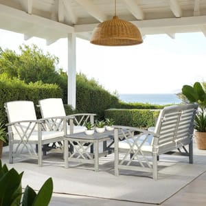 A 4-Person Acacia Conversation Set featuring a loveseat, two armchairs with white cushions, and a coffee table, set on an outdoor rug. It is situated under a covered patio with a wicker light fixture and overlooks lush greenery and a distant ocean view.