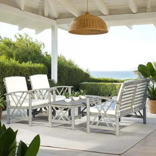 A 4-Person Acacia Conversation Set featuring a loveseat, two armchairs with white cushions, and a coffee table, set on an outdoor rug. It is situated under a covered patio with a wicker light fixture and overlooks lush greenery and a distant ocean view.