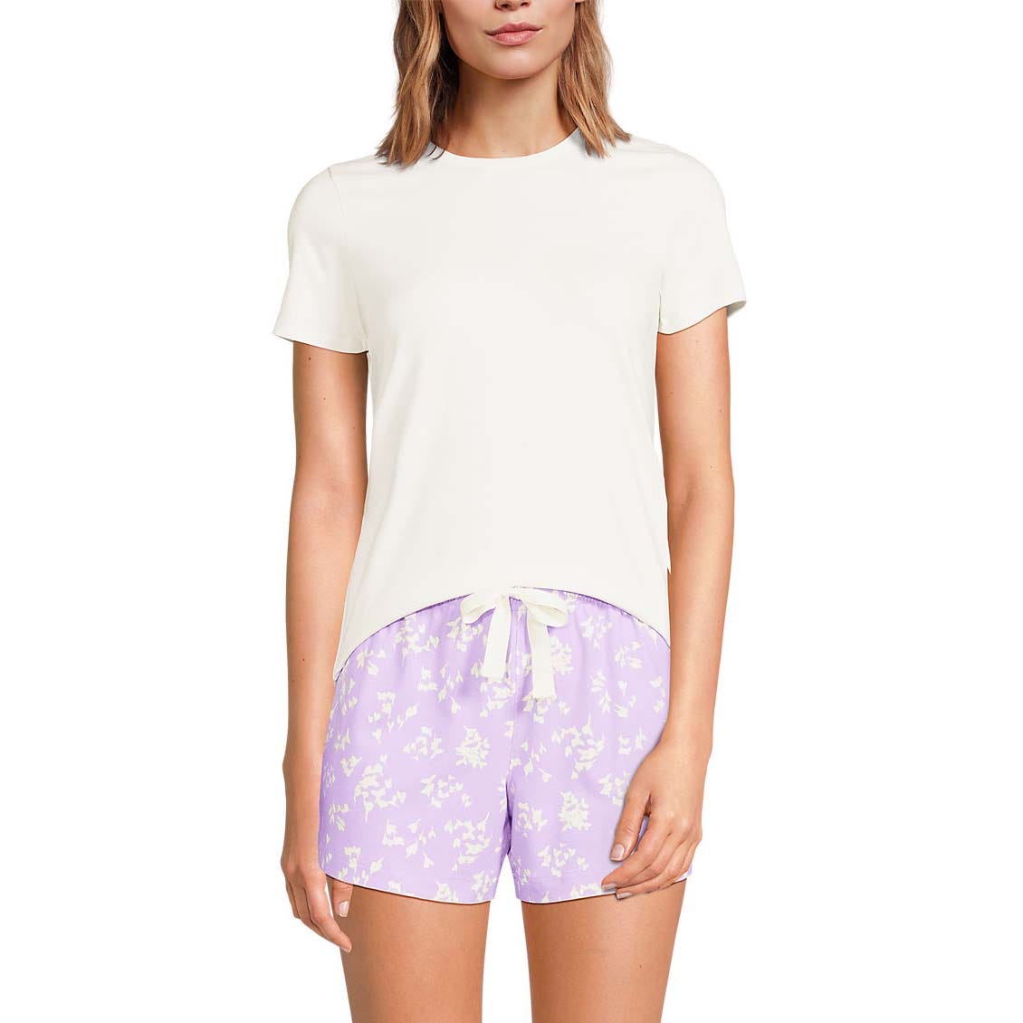 A woman is wearing a white short-sleeve T-shirt paired with lilac-colored shorts featuring a white floral pattern and a drawstring waist.