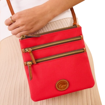 A red crossbody bag with three front zippered pockets and a brown leather strap is worn by a person dressed in a white top and beige skirt.