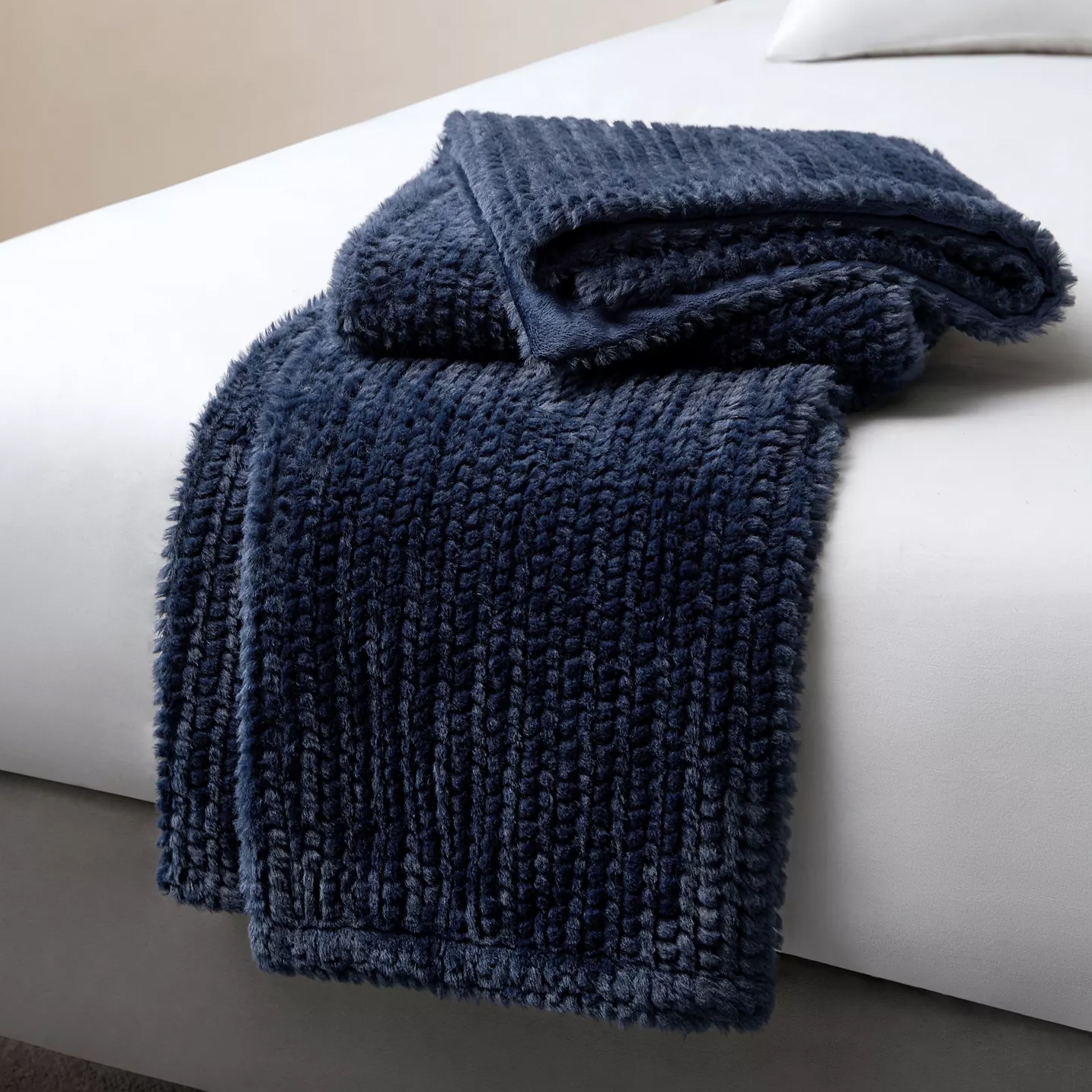 A plush, dark blue faux fur throw blanket is draped over the corner of a bed with a white sheet.