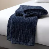 A plush, dark blue faux fur throw blanket is draped over the corner of a bed with a white sheet.