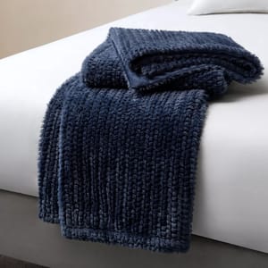 A plush, dark blue faux fur throw blanket is draped over the corner of a bed with a white sheet.